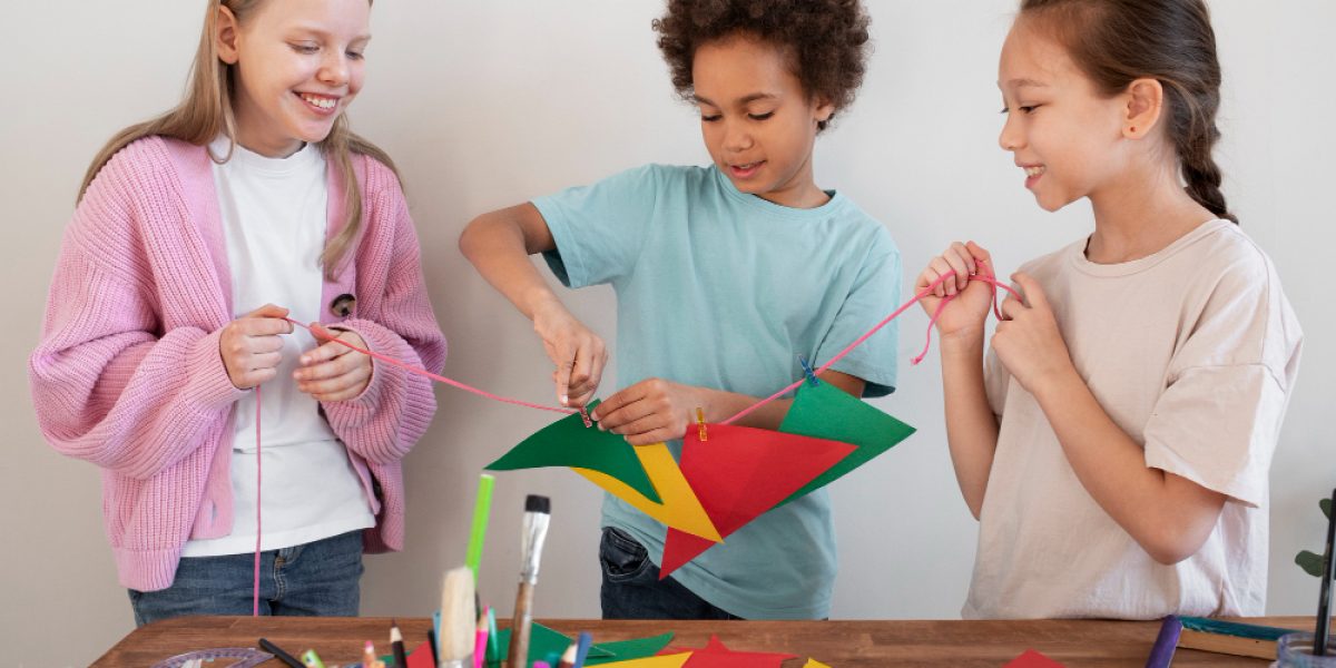 três crianças diversas em pé defronte a uma mesa brincam com materiais de artesanato.